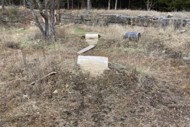 A natural landscape with sparse vegetation, featuring a gravel pathway leading toward two concrete blocks. Surrounding the area are bare trees, some with foliage, and a rocky ledge in the background. The sky is partly cloudy, suggesting an overcast day. Lehigh Portland Trails mountain bike trail.