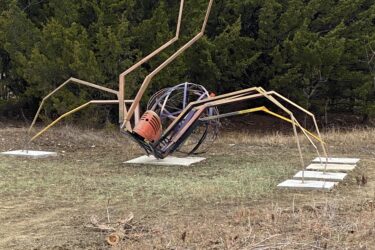 A large, artistic sculpture of a spider made from metal and other materials, featuring elongated legs and a rounded body, set against a backdrop of green trees. The sculpture is mounted on a grassy area with concrete bases. Lehigh Portland Trails mountain bike trail.