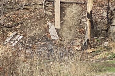 A weathered, sloped dirt area with exposed rock and some fallen branches, featuring a makeshift wooden platform extending outwards. In the foreground, a cylindrical concrete object sits partially buried in the ground, surrounded by sparse grasses and a few distant trees. The scene is set in a wooded environment with bare branches, suggesting a cool season. Lehigh Portland Trails mountain bike trail.