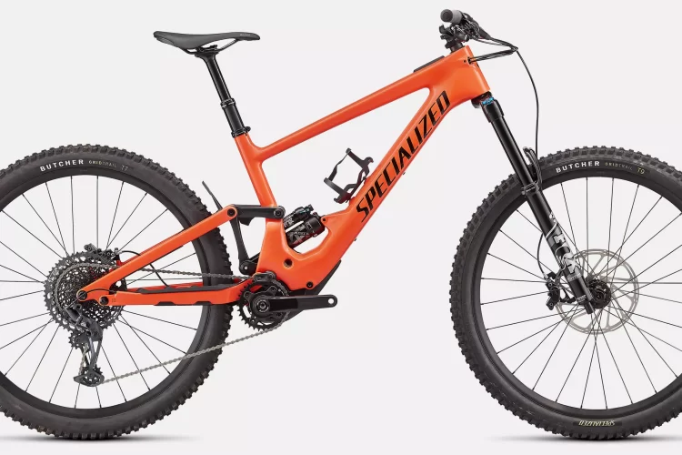 A bright orange mountain bike with a robust frame and large, treaded tires. The bike features a modern design with a prominent brand logo, a dual-suspension system, and a gear setup visible on the rear wheel. The handlebars are equipped with grips and a brake lever, showcasing its readiness for rugged terrains.