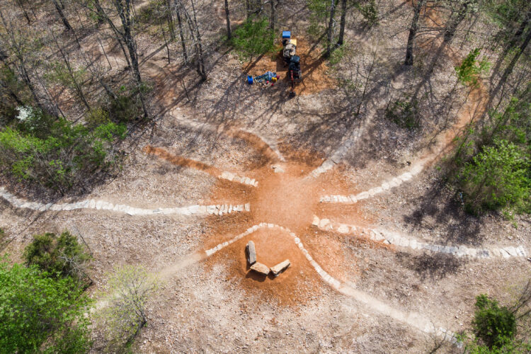 Aerial view of a woodland area featuring a series of dirt paths radiating outward from a central circular hub made of stone. In the background, a small campsite setup with a table and supplies can be seen among the trees, surrounded by a mix of greenery and bare ground.