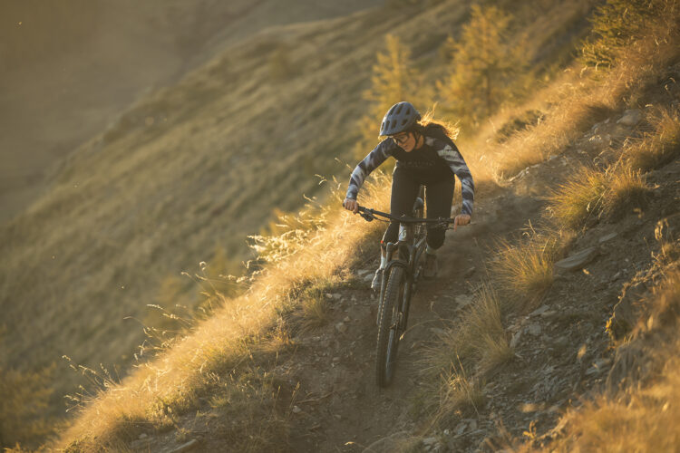 A person riding a mountain bike down a rocky trail during golden hour, surrounded by tall grass and a scenic landscape. The biker is focused and wearing a helmet and protective gear, with sunlight illuminating the environment.