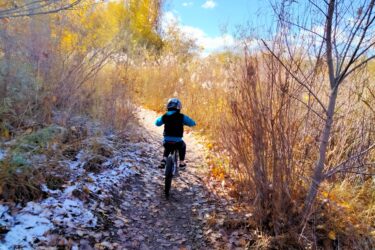 A child riding a bicycle along a narrow trail surrounded by autumn foliage and sparse, snowy patches. The scene features golden and rusty-colored leaves, with a bright blue sky peeking through the trees. Germania Park mountain bike trail.