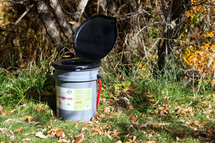 A portable camping toilet, known as a "Luggable Loo," positioned on grassy ground surrounded by autumn leaves and trees. The lid is open, revealing the seating area of the toilet.