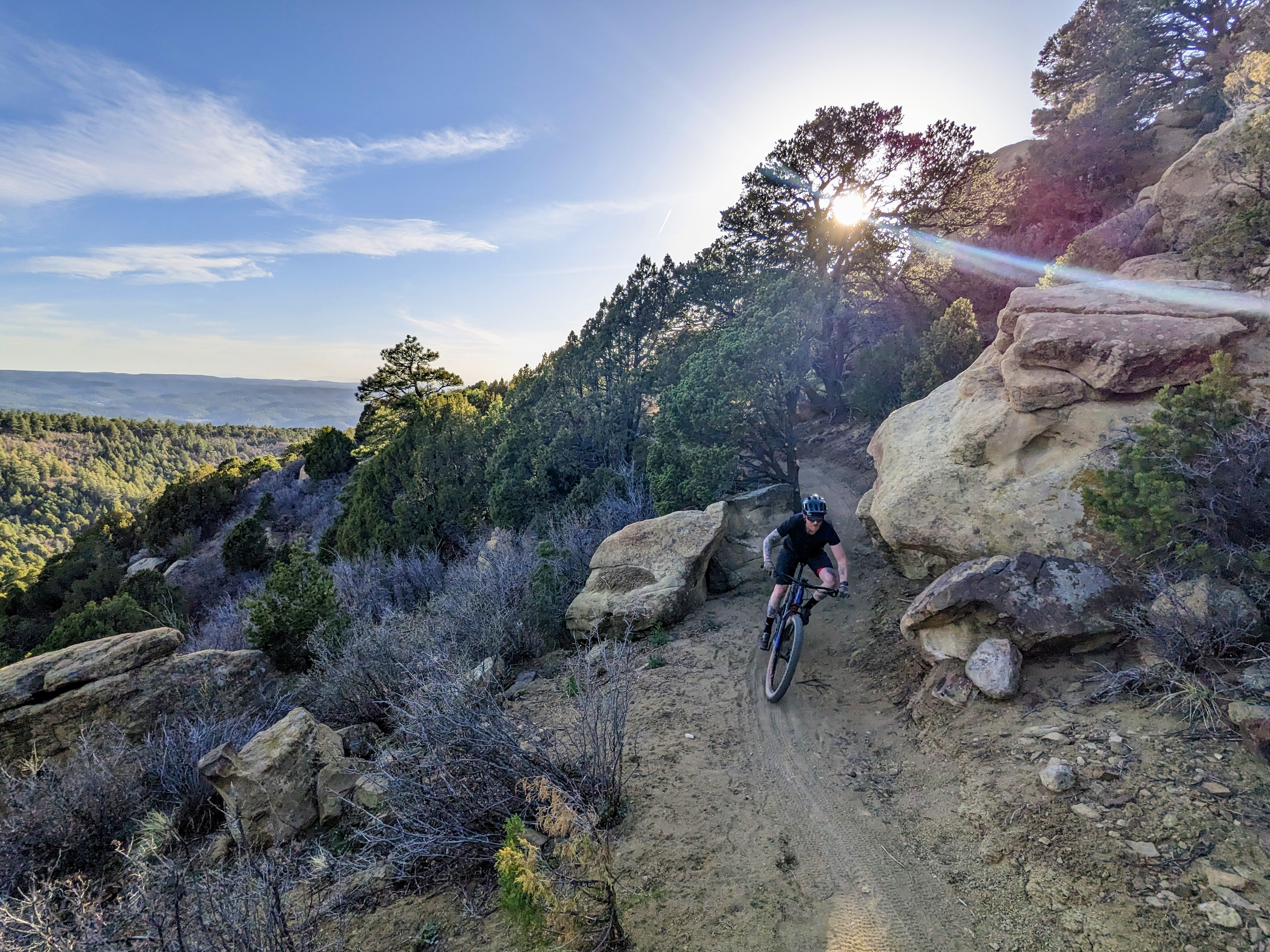A mountain biker navigating a rocky trail surrounded by trees, with the sun setting in the background, casting rays of light through the foliage. The landscape features a mix of dirt path and large boulders, showcasing a scenic outdoor adventure. Fishers Peak State Park mountain bike trail.