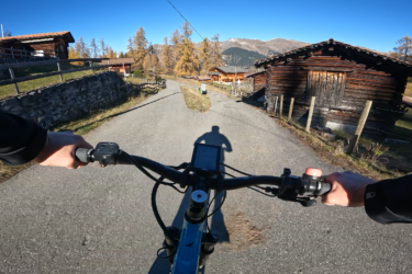 A cyclist's perspective while riding on a winding road surrounded by alpine scenery, with wooden chalets and autumn foliage in the background under a clear blue sky. The handlebars and shadow of the cyclist are visible in the foreground. Arosa to Chur mountain bike trail.