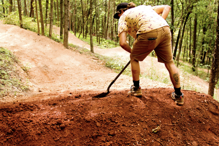 A person in a patterned shirt and shorts is using a shovel to work on a dirt jump in a forested area. The background features winding dirt paths and trees, suggesting an outdoor setting for cycling or biking activities.