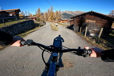 A view from the handlebars of a mountain bike, showing a gravel path winding through a scenic village with wooden cabins and autumn foliage. The sun is shining brightly in a clear blue sky, casting a shadow of the cyclist on the ground. Arosa to Chur mountain bike trail.