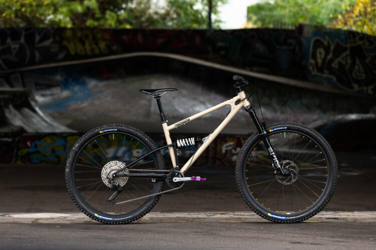 A modern mountain bike with a beige frame, black and yellow wheels, and a rear suspension system, parked in front of a graffiti-covered skatepark ramp.