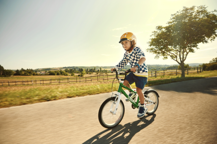 A child wearing a helmet rides a green bicycle along a sunny rural road, surrounded by fields and trees. The child is dressed in a checkered sweatshirt and shorts, focused on riding as they enjoy a clear blue sky in the background.