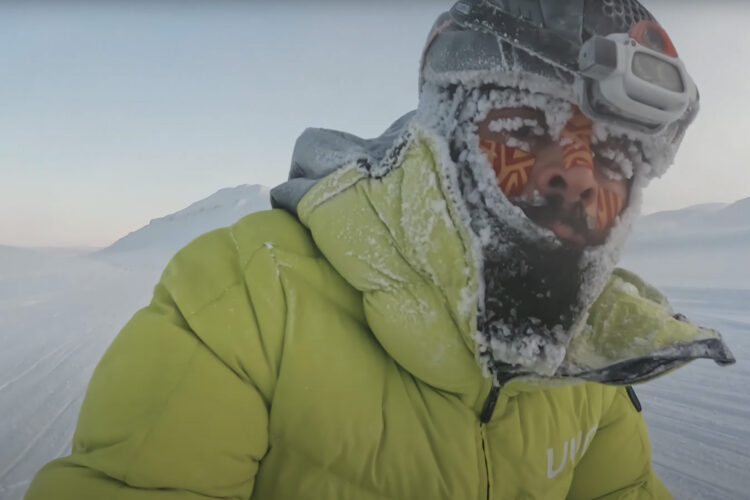 A person wearing a bright green puffy jacket and a headlamp stands in a snowy landscape, with frost covering their face and gear. The background shows a cold, mountainous terrain under a clear blue sky, emphasizing the extreme cold environment.