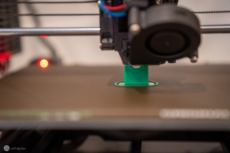 A close-up view of a 3D printer nozzle printing a small, green plastic object on the build plate. The printer features a sleek design with visible wiring and lights, indicating it's in operation.