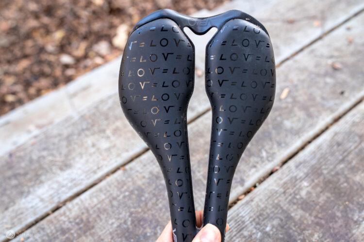 A close-up image of a bicycle seat with a unique design featuring the word "LOVE" repeatedly in various orientations. The seat is held by a hand, with a wooden surface and blurred background of leaves, suggesting an outdoor setting.