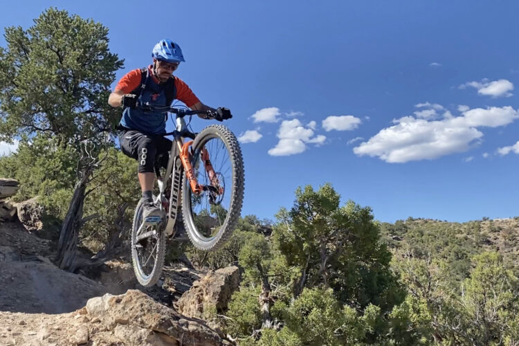 A mountain biker performs a jump on rocky terrain, showcasing a mid-air maneuver with one wheel elevated off the ground. The setting features a blue sky with white clouds and surrounding green trees, emphasizing an outdoor adventure environment.