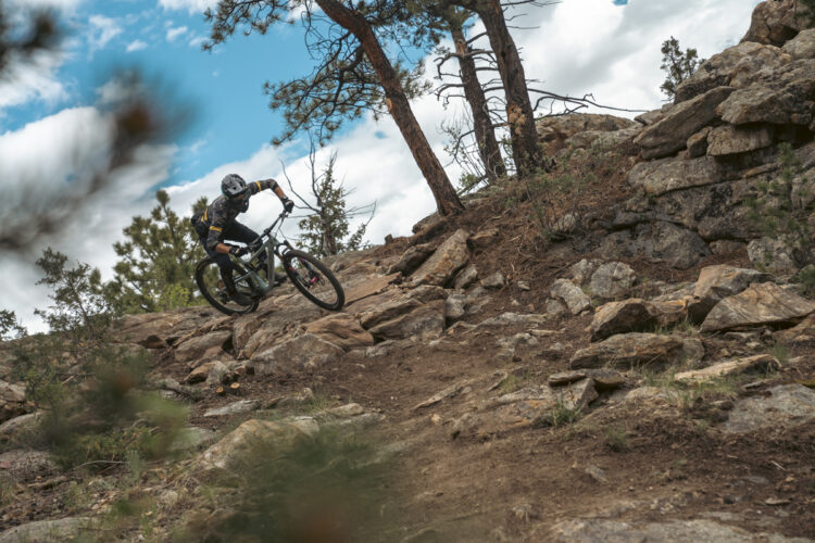 A mountain biker navigating a rocky trail surrounded by trees, showcasing skill and agility while descending a hillside under a cloudy sky.