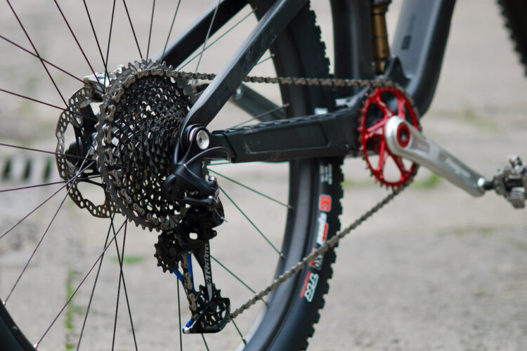Close-up view of a bicycle's rear wheel drivetrain, showcasing the cassette, derailleur, and chain. The components include a silver crank arm with a red chainring and a black cassette mounted to the hub. The bicycle tire is partially visible on the right side of the image. The background features a blurred concrete surface.