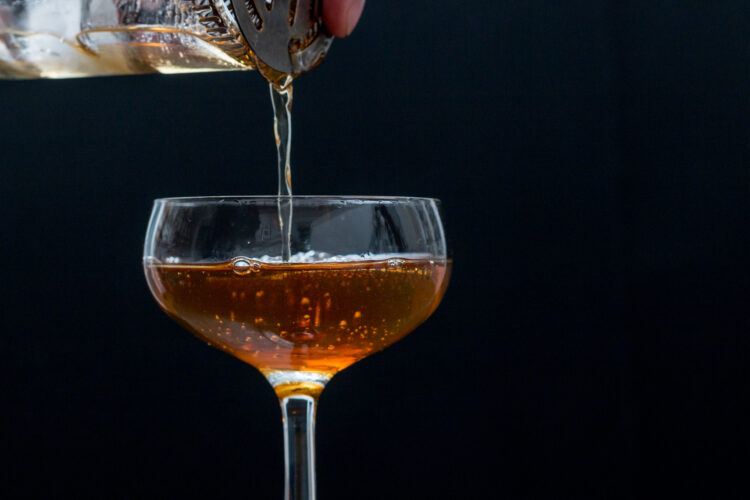 A cocktail being poured from a strainer into a stylish coupe glass, filled with a brownish beverage, against a dark background. The image captures the pouring action, highlighting the bubbles and clarity of the drink.