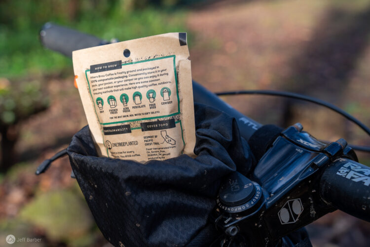 A close-up image of a coffee packaging labeled "Hiker's Brew Coffee" resting in the pocket of a bicycle's handlebar bag. The packaging features brewing instructions for different methods such as French Press and Pour Over, along with details highlighting sustainability. The blurred background shows natural terrain, hinting at an outdoor setting.