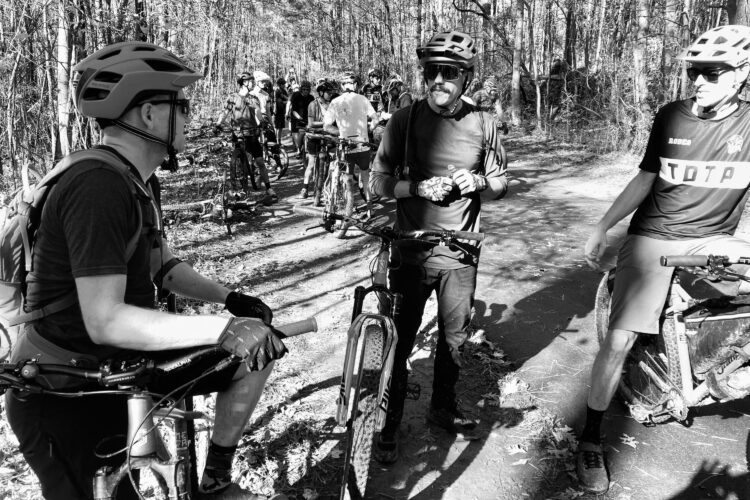 A black and white image of a group of mountain bikers gathered on a trail surrounded by trees. Three cyclists are prominently featured in the foreground; one is sitting on a bike, while the others stand beside their bikes engaged in conversation. The scene captures a moment of camaraderie among cyclists in a natural outdoor setting.