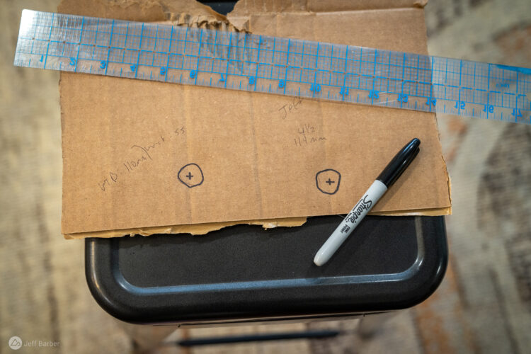 A close-up image of a piece of cardboard with handwritten measurements and markings. The cardboard is laid over a black surface, with a metallic ruler positioned above it. Two circular marks with a cross inside are visible on the cardboard, along with some scribbled notes indicating measurements, including "4½ 11mm." A black Sharpie marker is also partially visible in the foreground.