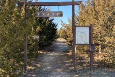 A wooden trail sign marked "Trail" stands at the entrance of a pathway surrounded by greenery, with a map of Cheney State Park displayed on a post nearby. The path is clear and leads into a wooded area under a bright blue sky. Cheney Lake Main Trail mountain bike trail.