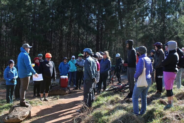 A group of participants gathered in a wooded area, listening to a speaker who is standing on a rock. The audience includes both men and women wearing cycling helmets and warm clothing, indicating a biking event or gathering. Bicycles are parked in the background, and the scene appears to be set in a natural outdoor environment, with tall trees surrounding the group.