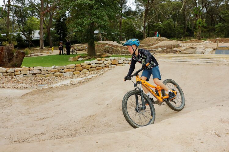 A young boy riding an orange mountain bike on a dirt track, surrounded by trees and rocky paths. In the background, two people are standing beside bikes in a green area. The setting appears to be a bike park or trail, showcasing a natural landscape.