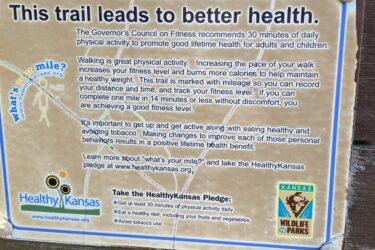 A sign on a wooden post with information about a health-focused trail. The text encourages walking as a form of physical activity to improve fitness and well-being. It includes recommendations from the Governor’s Council on Fitness, details on tracking walking distance, and a pledge promoting healthy habits such as exercising, eating well, and avoiding tobacco. The sign features logos for "Healthy Kansas" and "Kansas Wildlife & Parks." Cheney Lake Main Trail mountain bike trail.