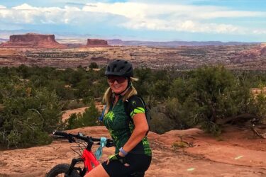 A woman wearing a cycling outfit and helmet poses with a red mountain bike on rocky terrain. The background features a scenic view of desert mesas under a partly cloudy sky. Ramblin' mountain bike trail.