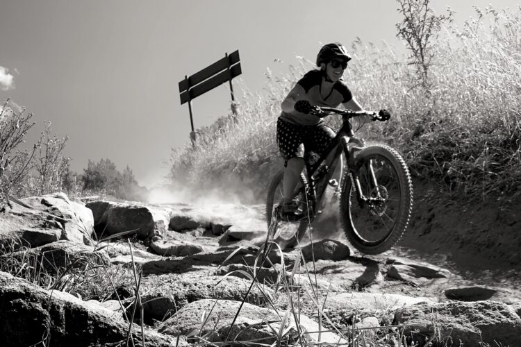 A mountain biker in a black and white setting rides over a rocky trail, kicking up dust as they navigate the uneven terrain. Tall grasses and a signpost can be seen in the background, suggesting an outdoor trail environment. The biker is wearing a helmet and sunglasses, and is captured mid-action.
