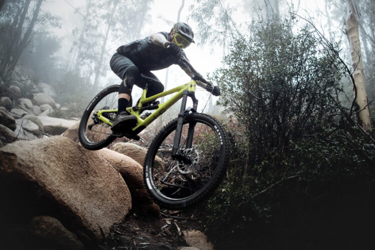 A mountain biker jumps over a large rock on a trail surrounded by trees, with fog creating a mysterious atmosphere. The biker is wearing a helmet and protective gear, and the mountain bike is bright yellow, contrasting with the earthy tones of the rocks and surrounding foliage.