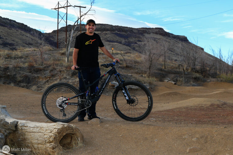 A person standing with a mountain bike in a rugged outdoor setting, featuring rolling terrain and rocky hills in the background. The individual is wearing a black t-shirt and jeans, smiling at the camera. Power lines are visible in the distance, and the sky is partly cloudy.