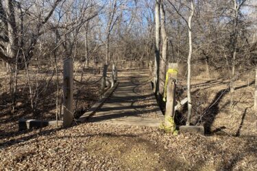 A wooden bridge surrounded by bare trees and fallen leaves, leading into a wooded area. Caution tape is visible on the posts at either end of the bridge. The scene is brightened by clear blue skies in the background. Cheney Lake Main Trail mountain bike trail.
