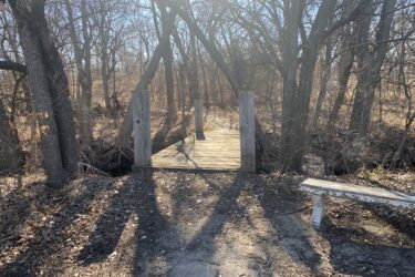 A wooden footbridge crosses a small creek, surrounded by barren trees and dry foliage in a wooded area. A weathered bench is positioned nearby, with soft sunlight filtering through the trees. Cheney Lake Main Trail mountain bike trail.