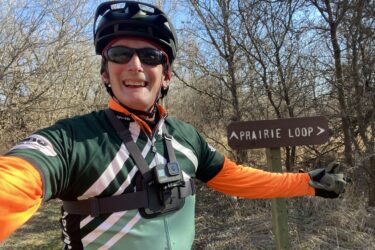 A smiling person in cycling gear, wearing a helmet and sunglasses, poses for a selfie while gesturing towards a brown sign marked "Prairie Loop." The background features bare trees and open grassland under a blue sky. The cyclist is equipped with a camera mounted on their chest. Cheney Lake Main Trail mountain bike trail.