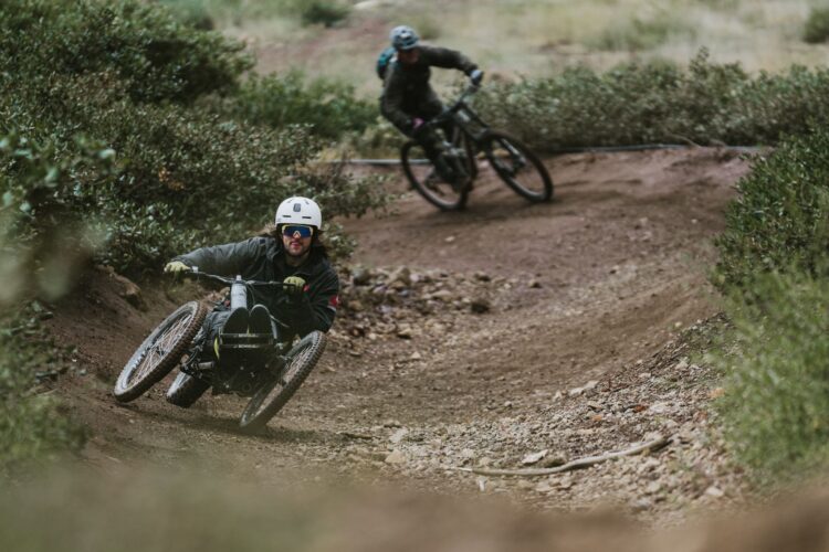 A person riding a handcycle navigates a dirt trail, leaning into a turn. Another cyclist follows closely behind on a traditional mountain bike, surrounded by greenery and a rugged terrain.