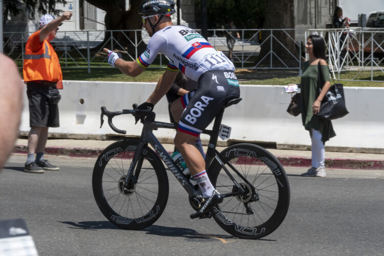 A cyclist wearing a white and black race jersey with colorful stripes rides a sleek black racing bike on a sunny street. In the background, a man in an orange vest raises his fist in encouragement, while a woman in a green top and white pants stands nearby, holding a bag. Barriers and spectators line the road, suggesting a cycling event or race is taking place.