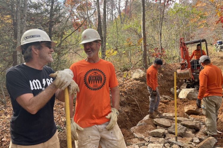 A group of men in hard hats and gloves are working on a trail construction project in a wooded area during autumn. Two men are smiling and chatting with a yellow tool between them, while others operate machinery and handle stones in the background. The trees are partially bare, showcasing fall foliage.