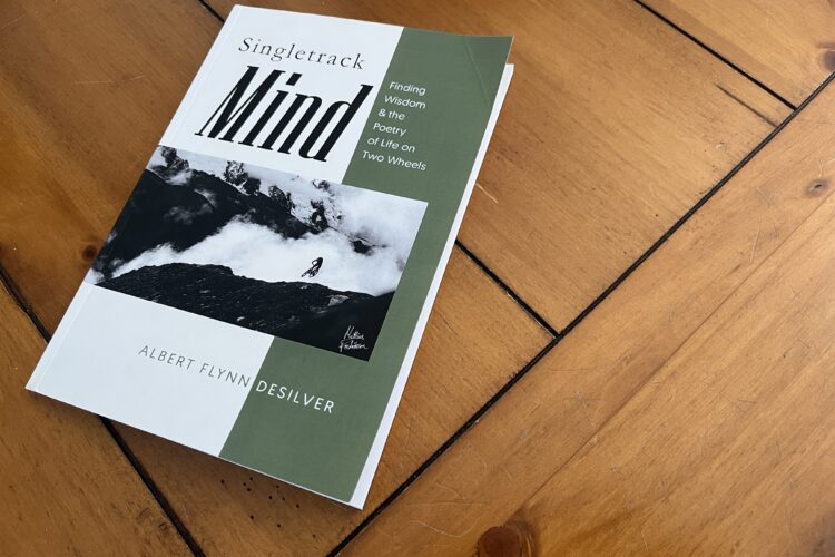 A book titled "Singletrack Mind: Finding Wisdom &amp; the Poetry of Life on Two Wheels" by Albert Flynn Desilver, resting on a wooden table. The cover features a black-and-white photograph of a mountain biker amidst a misty landscape, along with the author's name.