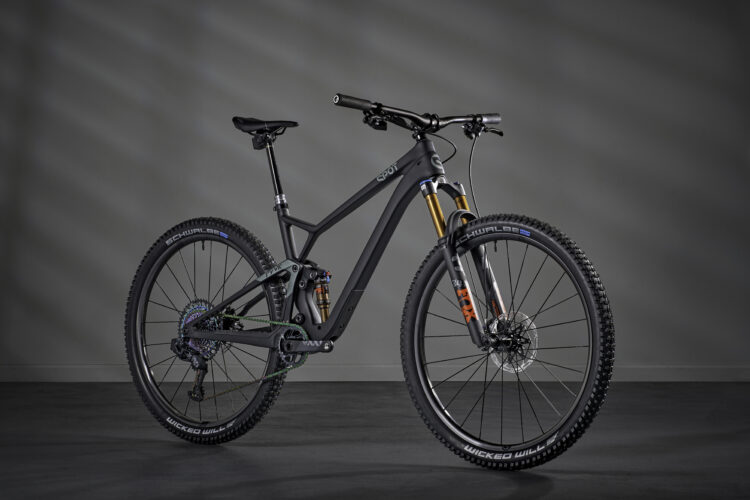 A sleek, modern mountain bike featuring a matte black frame, front suspension, and wide, knobby tires. The bike is positioned against a gray backdrop, showcasing its high-performance components, including a gold fork and colorful gear mechanism.