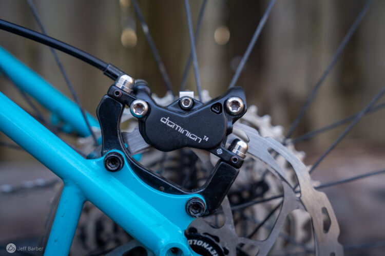 Close-up image of a bicycle's brake system, featuring a black brake caliper labeled "dominion T4" attached to a turquoise frame. The brake lines and part of the rotor are visible in the background, showcasing the mechanical components of the bike.