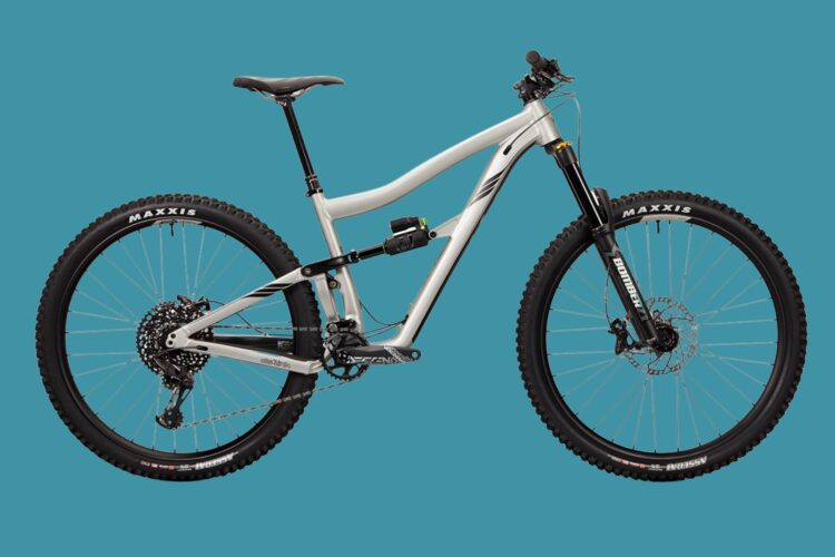 A modern mountain bike with a lightweight frame, featuring dual suspension, a dropper seat post, and rugged tires labeled "Maxxis." The bike is displayed against a solid turquoise background.