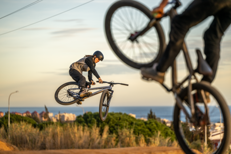 A young rider performs a mid-air trick on a mountain bike, with a scenic backdrop of ocean and hills. Another cyclist is seen in the foreground, showcasing their bike's wheel, as the sun sets in the background, casting a warm glow over the scene.