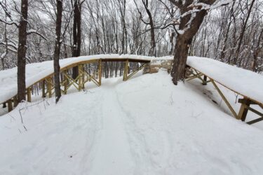 A snow-covered wooden bridge arches over a trail surrounded by bare trees. The path leads through a winter landscape, with snow blanketing the ground and branches. The scene conveys a peaceful, serene atmosphere typical of a snowy forest in winter. Monarch mountain bike trail.