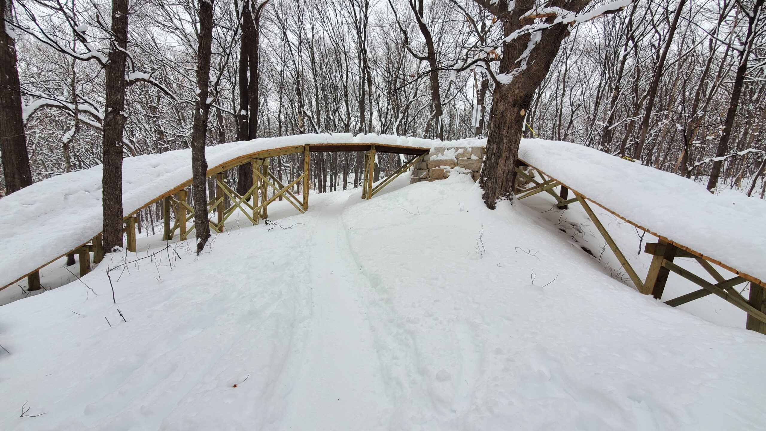 A snow-covered wooden bridge arches over a trail surrounded by bare trees. The path leads through a winter landscape, with snow blanketing the ground and branches. The scene conveys a peaceful, serene atmosphere typical of a snowy forest in winter. Monarch mountain bike trail.