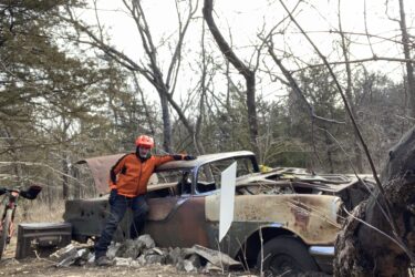 A person wearing an orange jacket and helmet leans against a rusted vintage car that is partially covered in foliage, surrounded by trees in a wooded area. A bicycle is nearby, and the setting appears to be an abandoned location with rocks scattered around the car. Cedar Niles Park mountain bike trail.