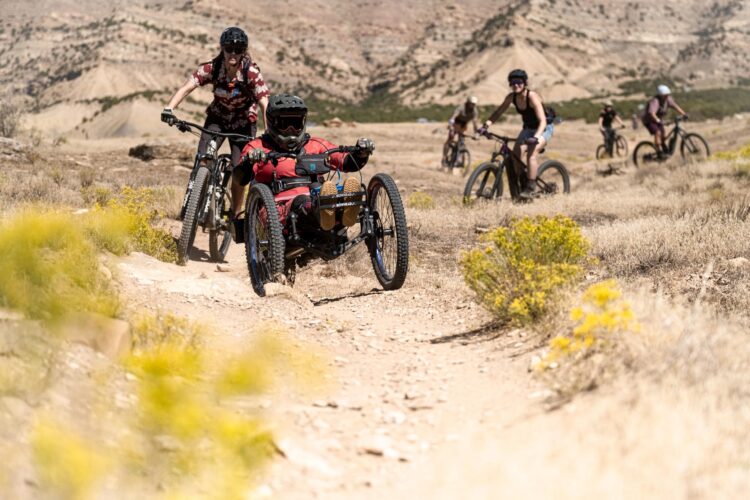 A group of mountain bikers traverses a rocky dirt trail in a desert landscape. One rider using a handcycle in a red jersey is being assisted by another cyclist, while additional bikers follow in the background. The scene captures the camaraderie and adventure of outdoor biking.
