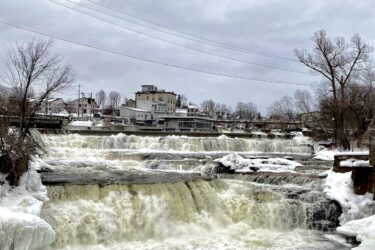 A scenic view of a waterfall cascading over ice-covered rocks, surrounded by trees and a cloudy sky. In the background, a few buildings are visible on the riverbank, adding a human element to the natural landscape. Almonte Riverside Trail mountain bike trail.
