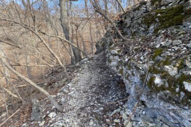 A narrow dirt trail winding along a rocky outcrop, surrounded by bare trees and scattered leaves. The scene is set in a wooded area during late fall or early winter, with blue skies visible through the branches. Cedar Niles Park mountain bike trail.