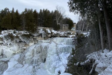A scenic view of frozen waterfalls surrounded by snow-covered trees, with a cloudy sky overhead. The ice formations cascade down the rocks, creating a beautiful winter landscape. Inglis falls West rock mountain bike trail.